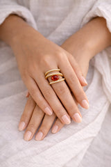 Close-up of hands wearing gold rings with a red gemstone on a light fabric background